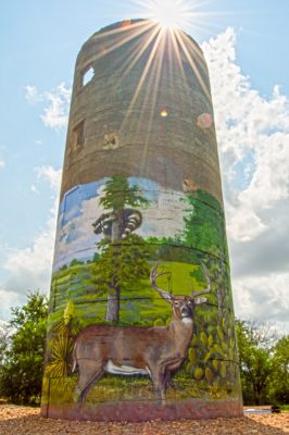 Wide-shot picture, with the camera pointing up from the base, of a tower with a mural painted by rafael acosta jr, and the sun shining just over the top of the tower, rafael acosta mural paintings