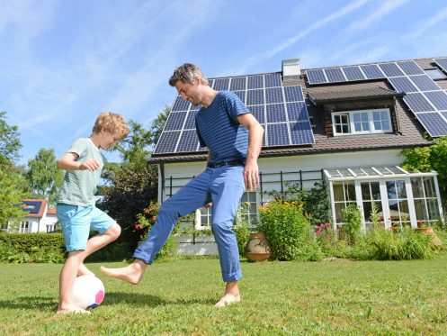 Father and son playing soccer on a sunny day with home with solar panels on roof in the background.
