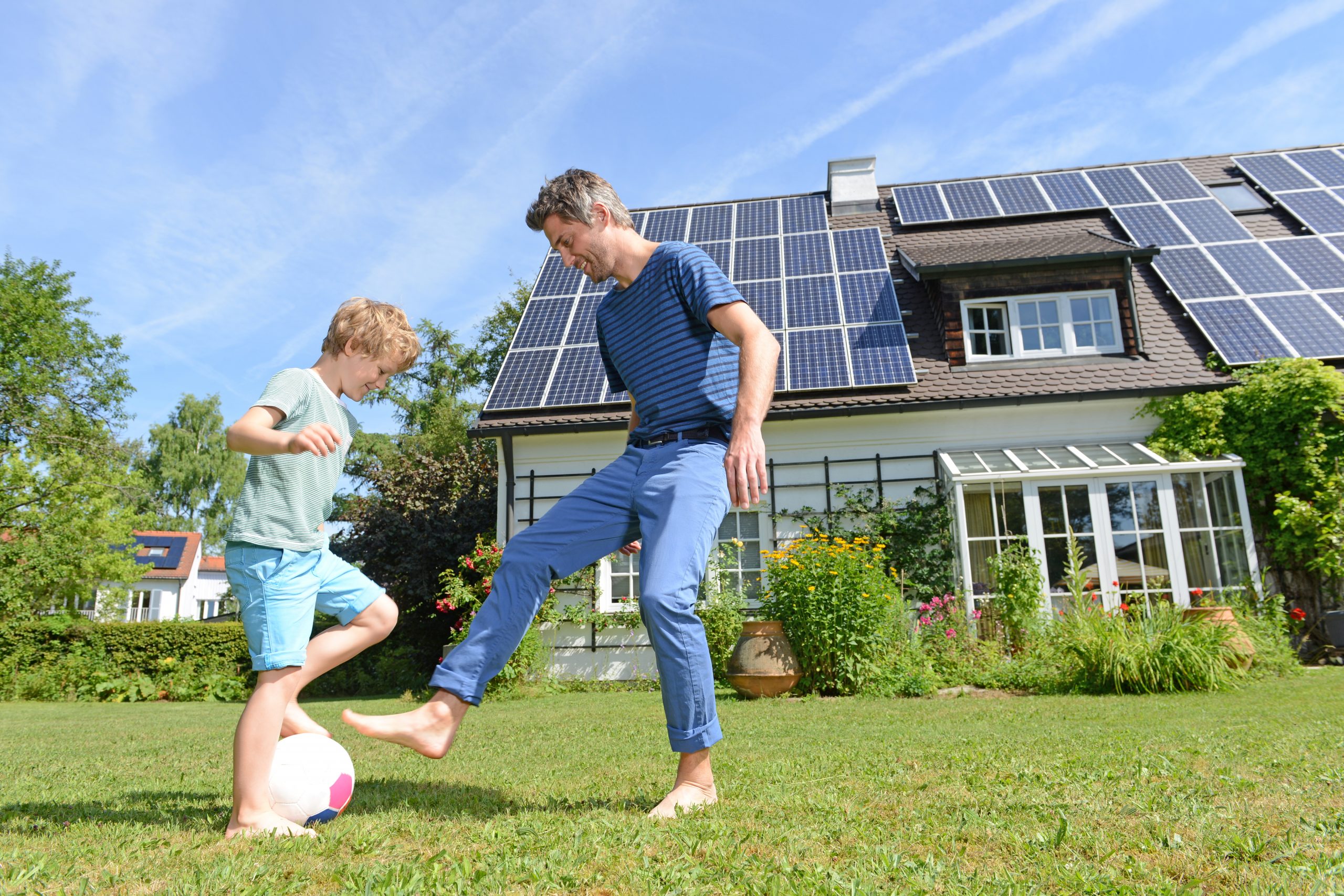 Father and son playing soccer on a sunny day with home with solar panels on roof in the background.