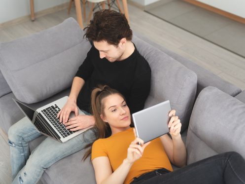 Couple on couch, using wi-fi signal in their home to connect to their wireless devices. Fiber and Wireless internet are both suitable for their wi-fi needs.