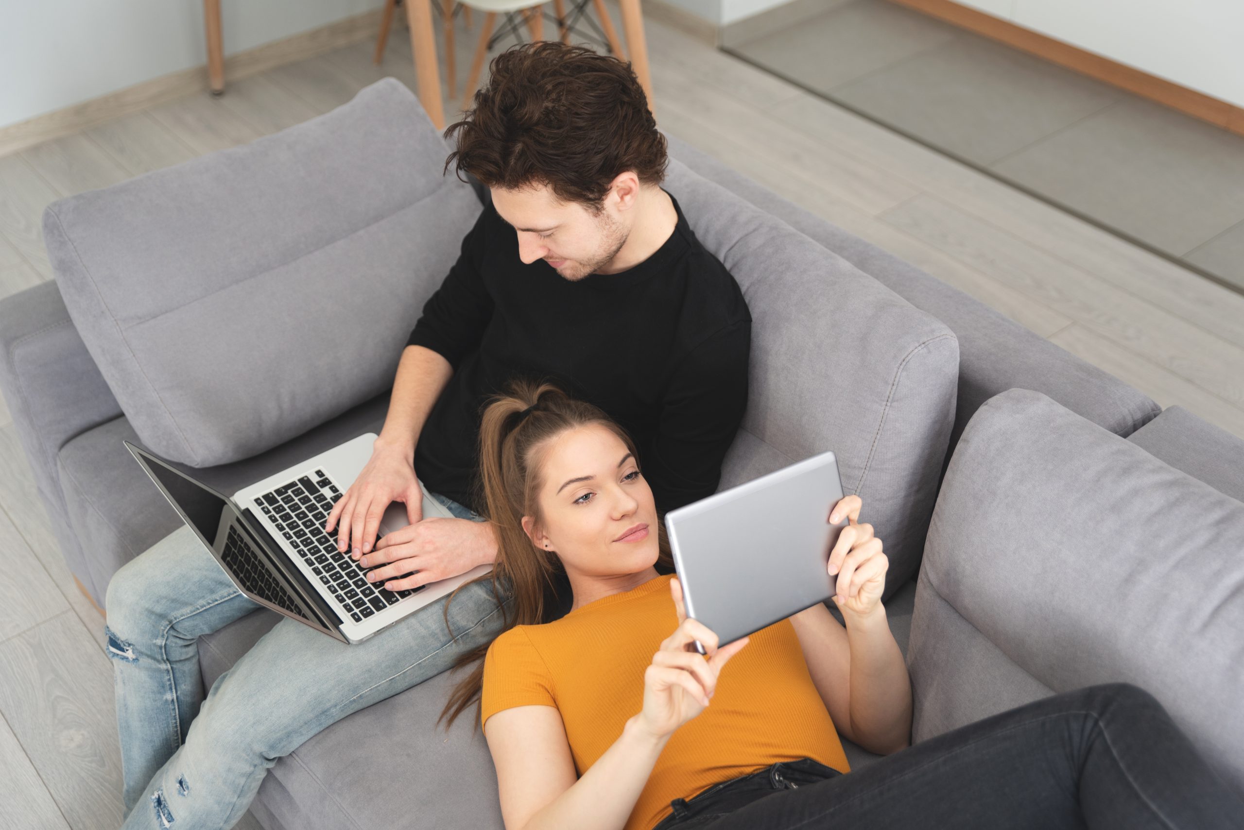 Couple on couch, using wi-fi signal in their home to connect to their wireless devices. Fiber and Wireless internet are both suitable for their wi-fi needs.